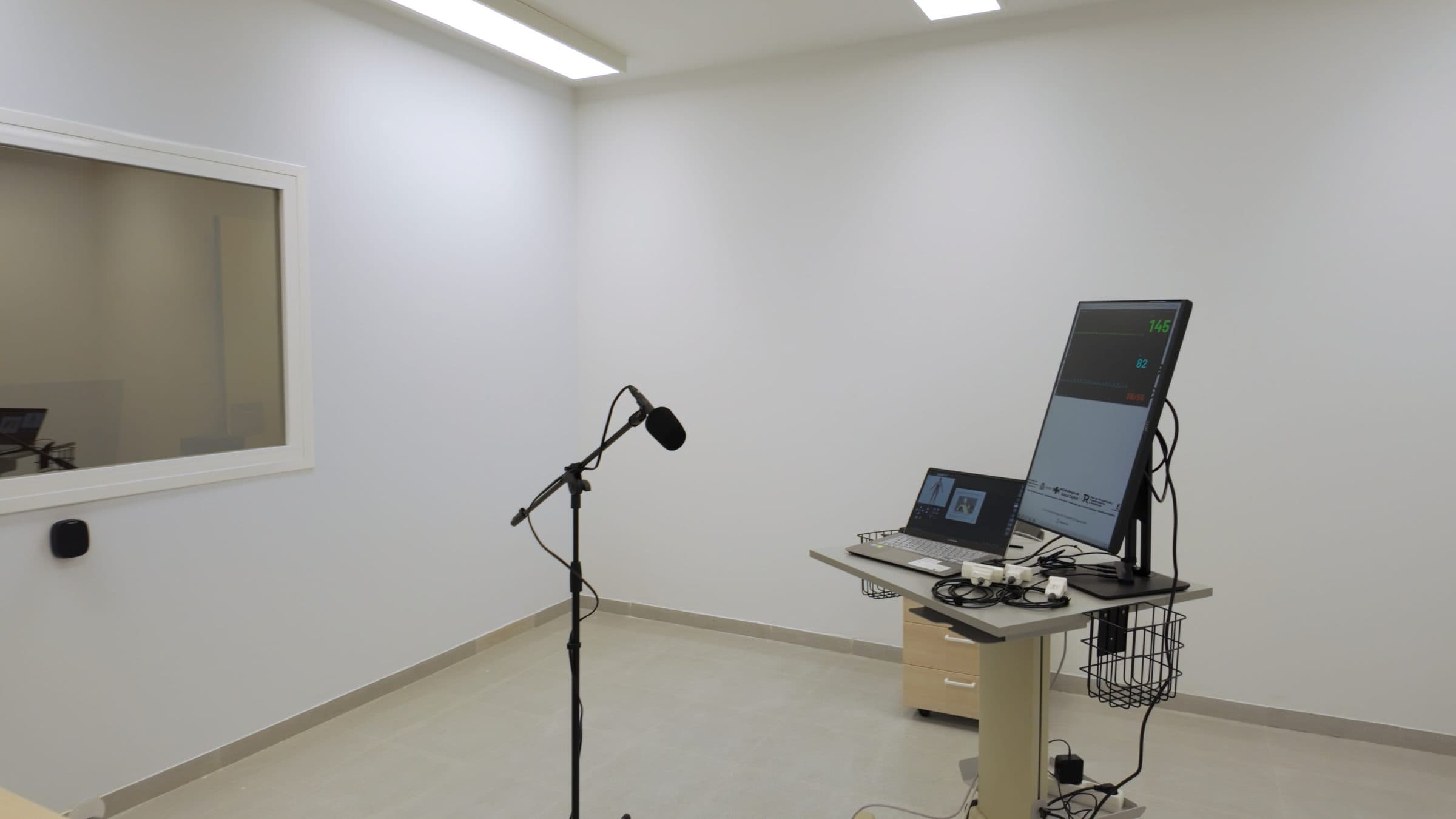 Aula de simulación clínica con el carro del monitor de constantes, el ordenador del simulador y un micrófono de ambiente sobre pie, frente a la ventana que da al puesto de control.