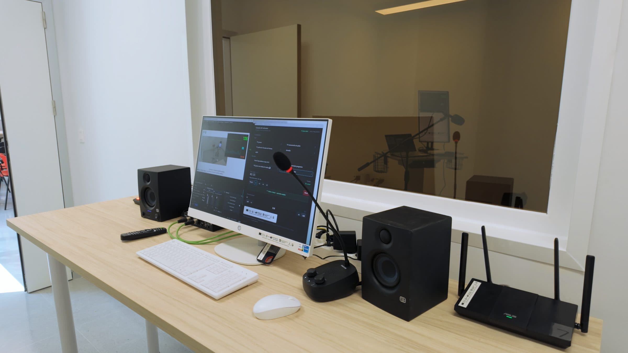 Puesto de control audiovisual con ordenador todo en uno, monitores de estudio, micrófono de intercomunicación y router, con la sala de práctica visible a través de la ventana.